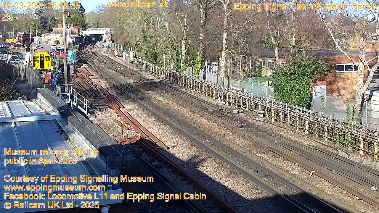 A sunny day at a train station shows multiple train tracks, a railway car, and buildings beyond a fence.