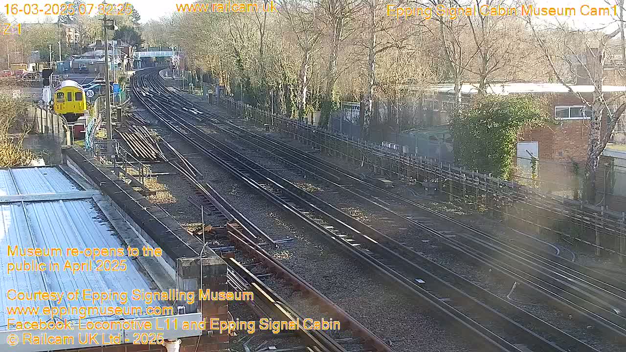 A sunny view of a railway station with multiple train tracks, a yellow train, and trees.