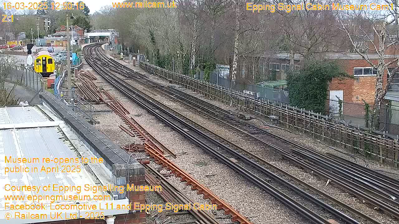 A high-angle view of multiple train tracks on a partly sunny day, with a yellow train car visible in the distance and leafless trees surrounding the railway.