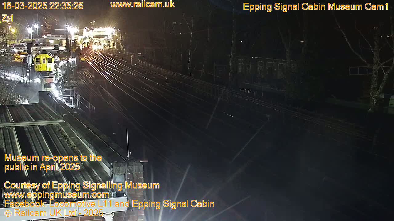A nighttime view of a train station with multiple train tracks and a yellow train partially visible, under artificial light.