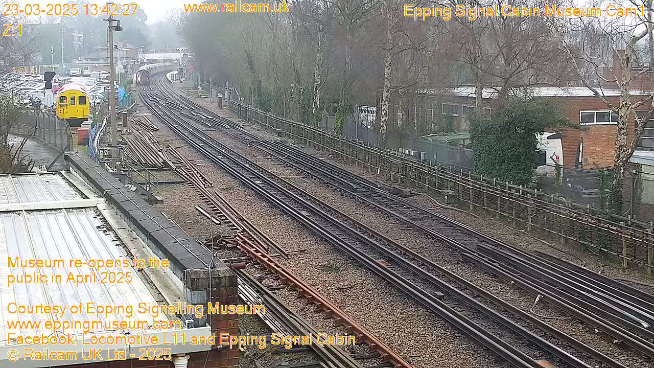 A yellow train is visible on a railway track, passing through a station on an overcast day.