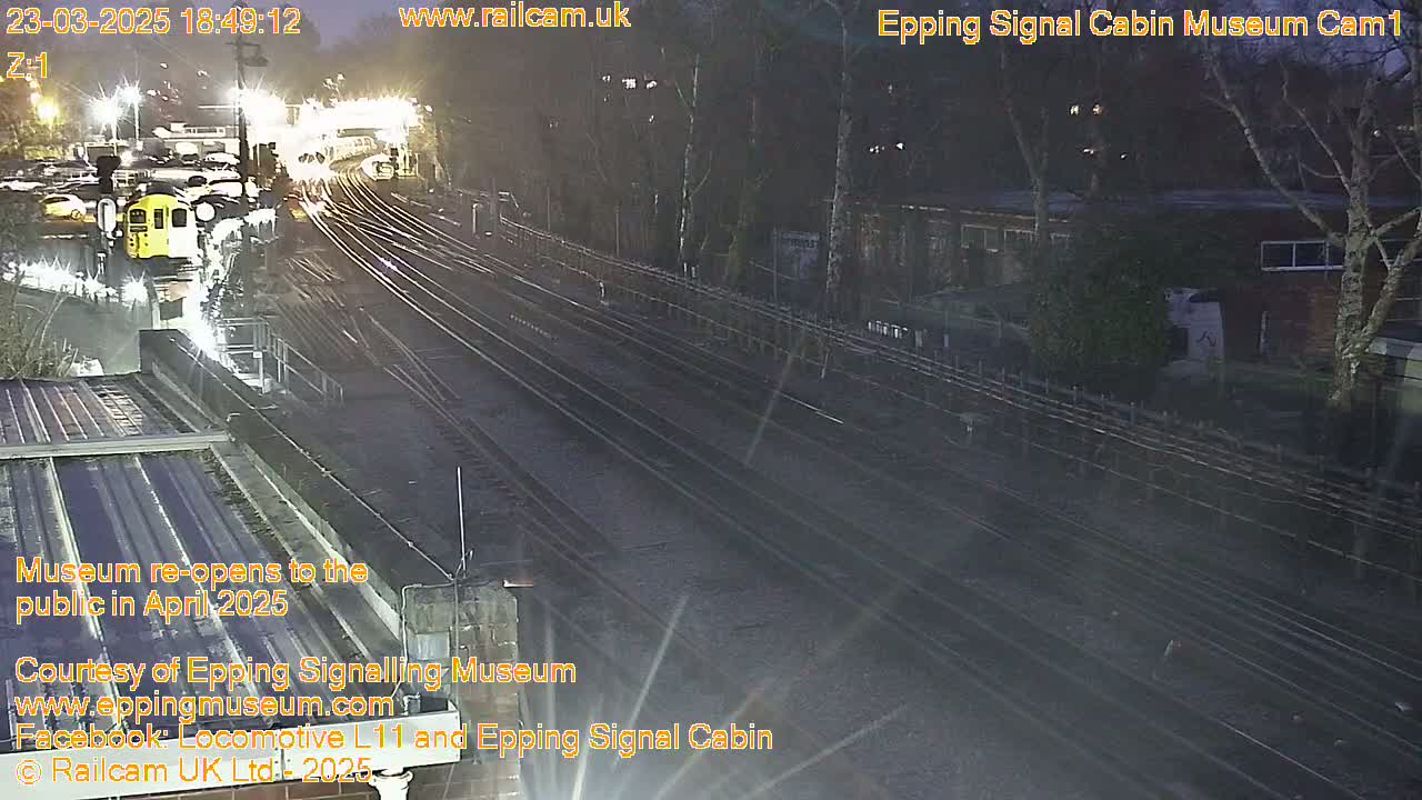 A nighttime view of train tracks with a train and several buildings visible, surrounded by trees under dark conditions.