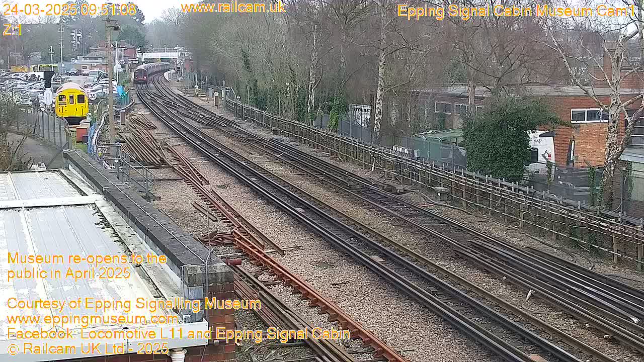 A high-angle, long shot on a partly cloudy day shows multiple train tracks with a yellow train car and a longer train in the distance, alongside a building and trees.