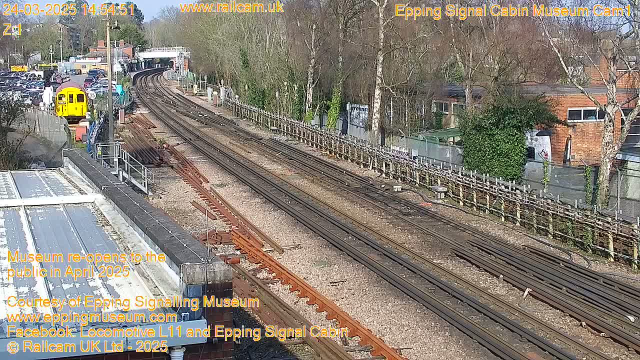 A sunny day at a train station shows multiple train tracks, a yellow train car, and a brick building beyond a fence.