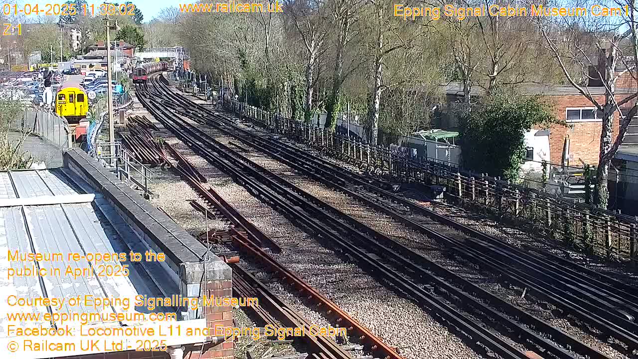 A sunny day at a railway yard shows multiple train tracks, a yellow train car, and a few parked cars near a wooded area.