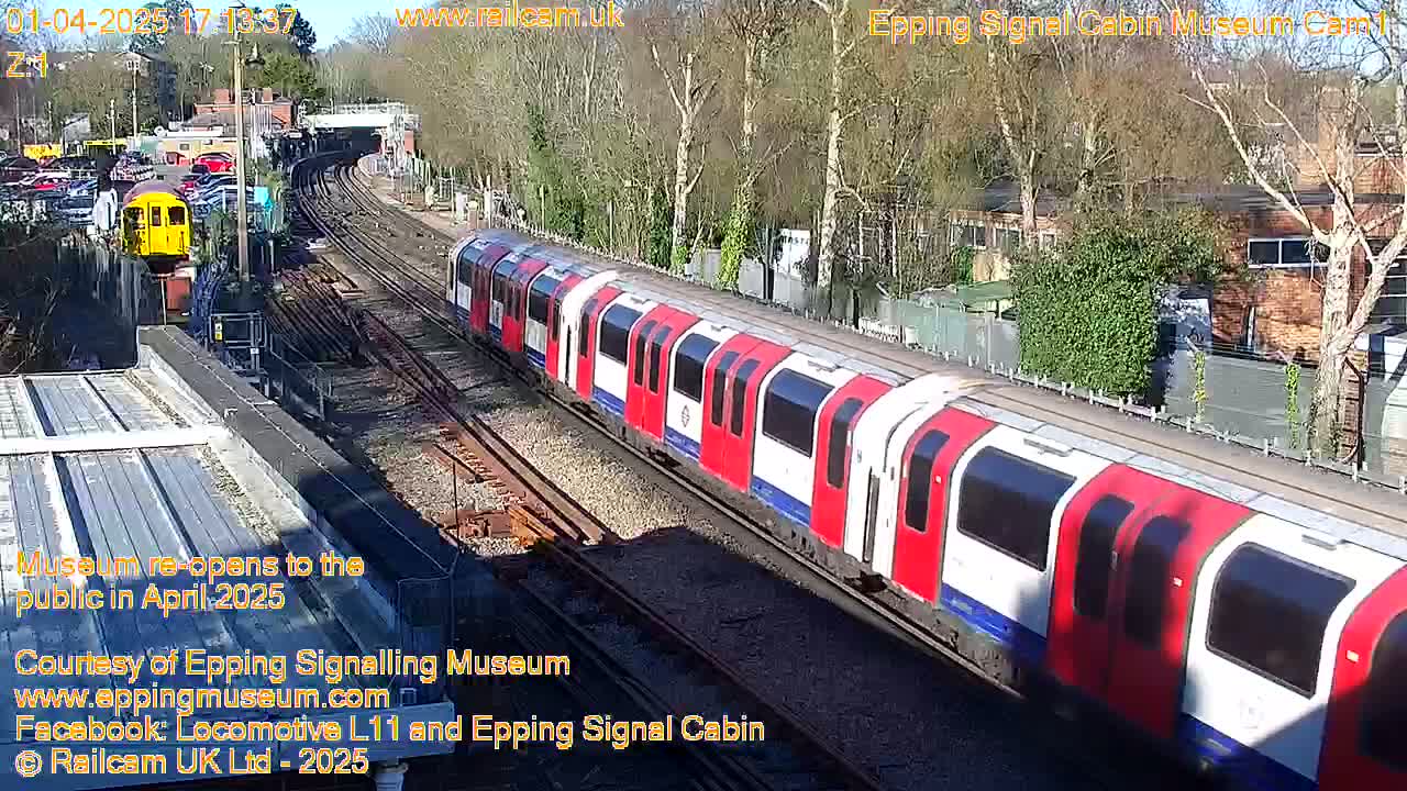 A long red and white train travels along train tracks past a yellow train and several parked cars under a sunny sky.