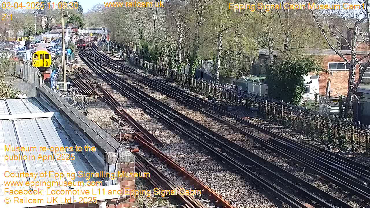 A sunny day at a railway yard shows multiple train tracks, a yellow train car, and a gray train engine partially obscured by trees and buildings.