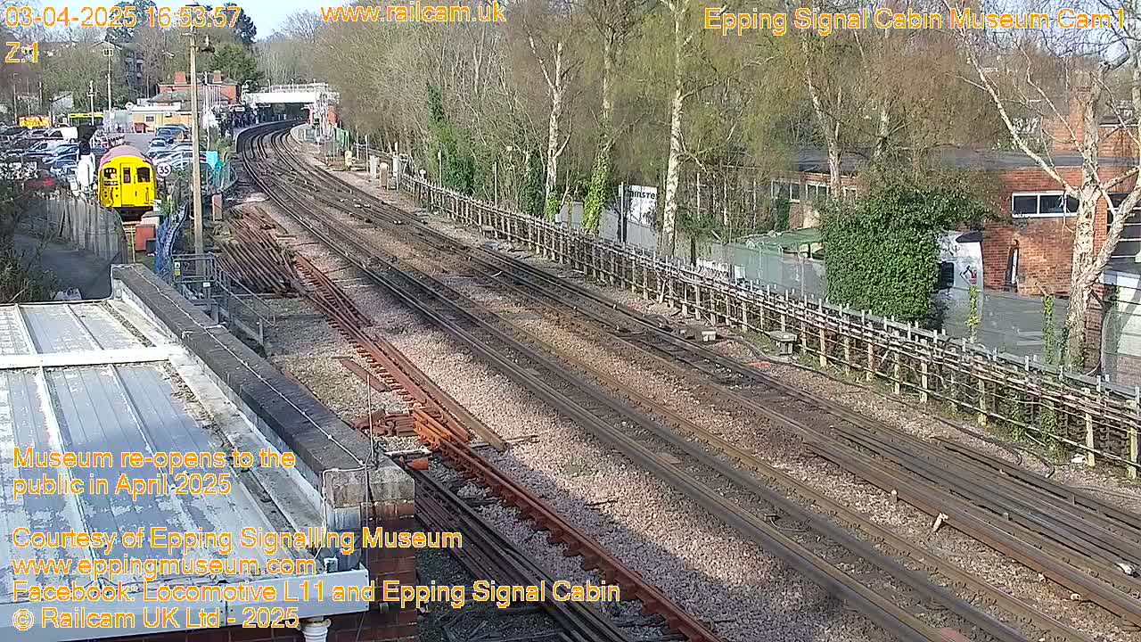 A yellow train is visible near a railway station on a sunny day, with multiple train tracks extending into the distance.