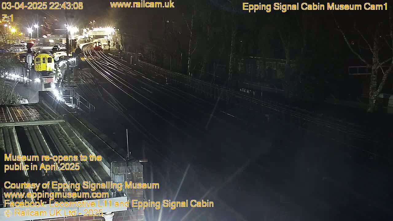A nighttime, overhead view shows a train station with multiple train tracks and a yellow train partially visible, surrounded by dark trees under artificial lighting.