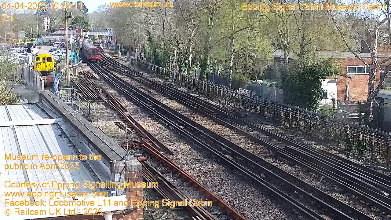 Two trains are visible on a sunny day on multiple railway tracks that run alongside a fenced area with trees and buildings.