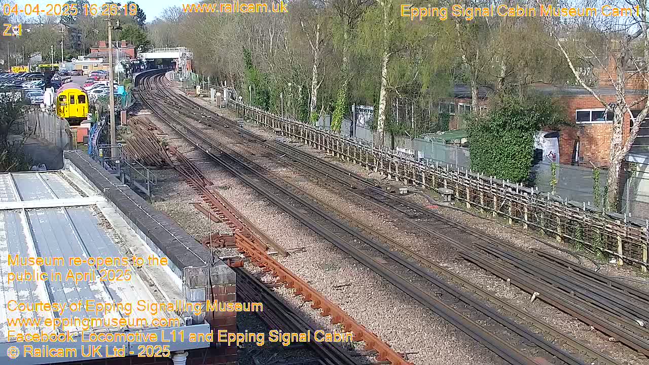 A sunny day reveals multiple train tracks extending into the distance, alongside a railway station with a yellow train car visible.
