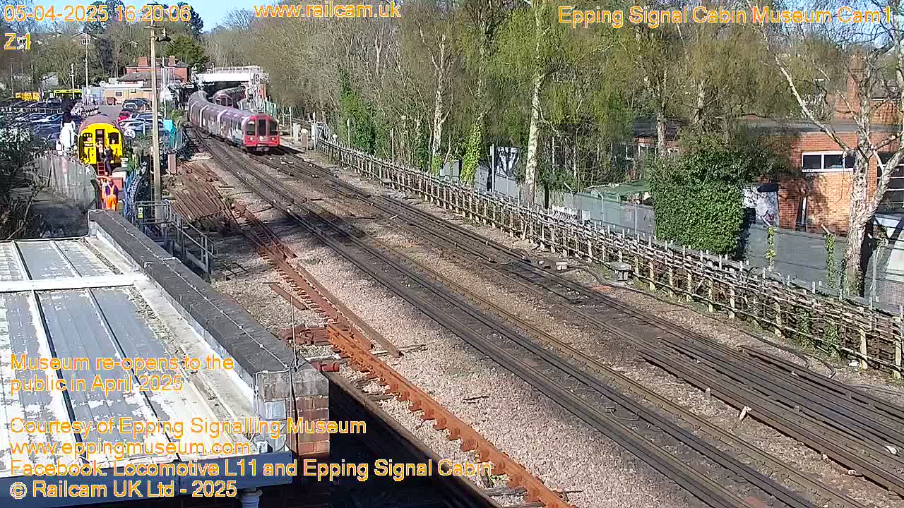 A sunny day at a railway station shows two trains on parallel tracks, one a longer passenger train and the other a shorter, possibly maintenance, vehicle; additional tracks and railway infrastructure are visible, alongside a fenced area and buildings in the background.