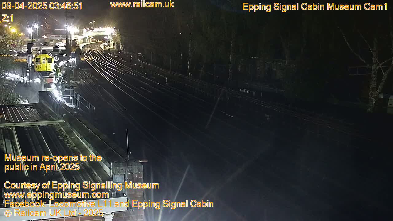 A nighttime, overhead view shows a train stopped on dark railroad tracks next to a dimly lit platform.