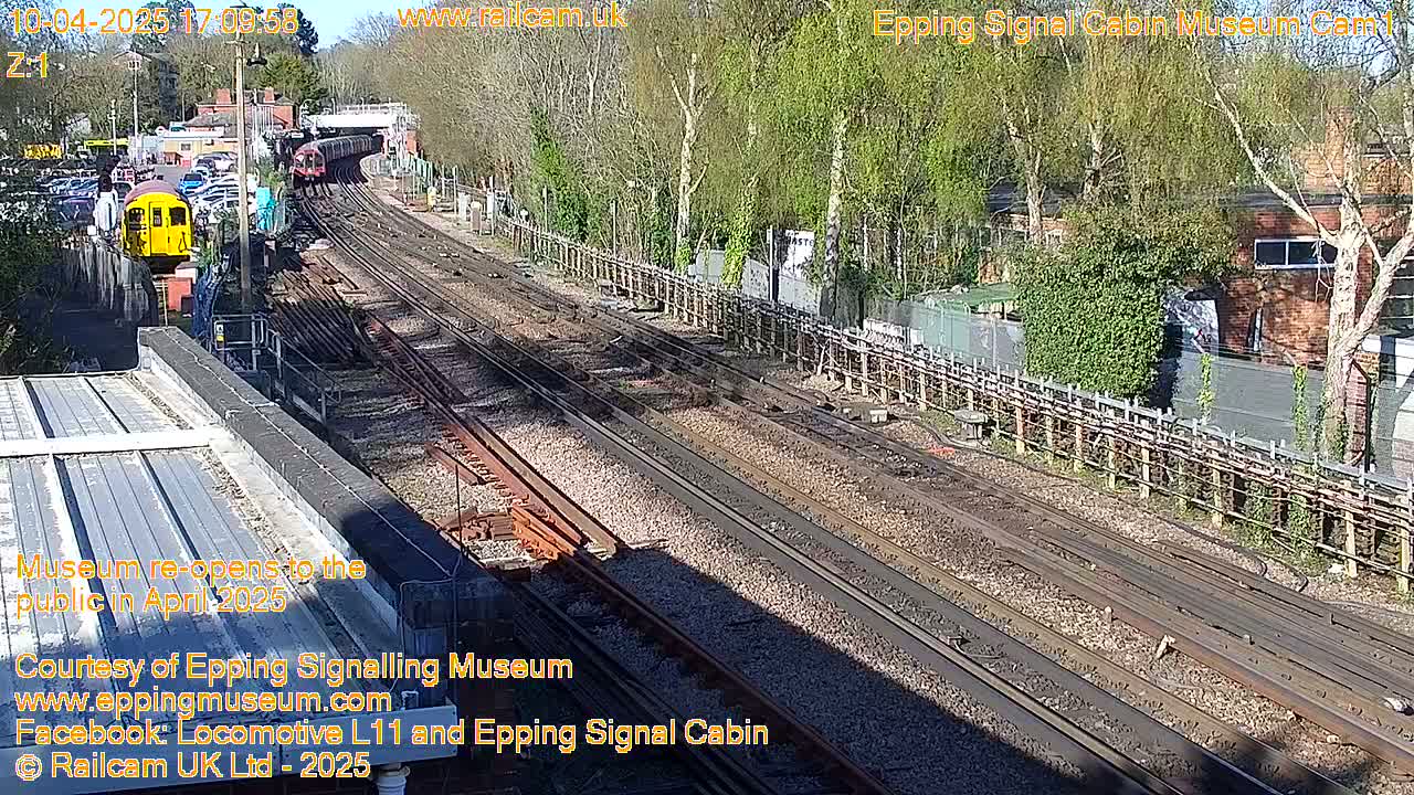 A sunny day at a train station shows multiple train tracks with a yellow train car and a longer train partially visible in the distance.