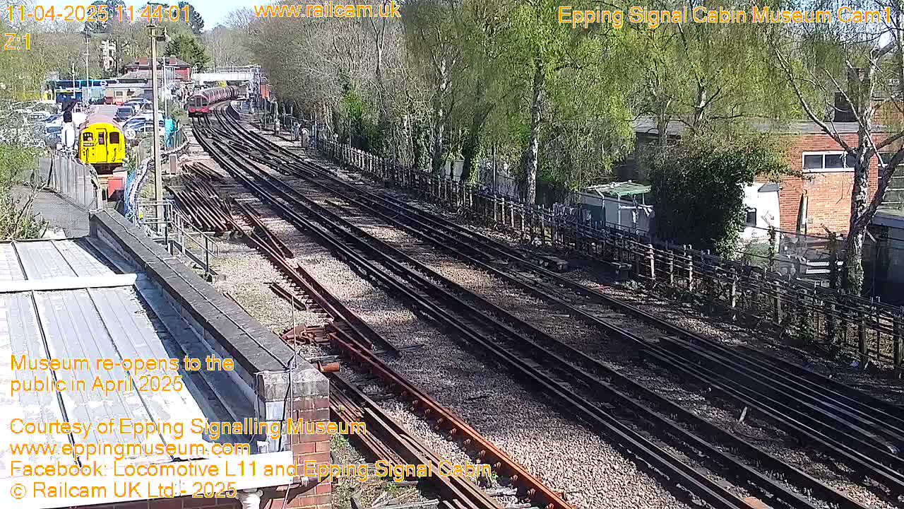 A sunny day at a railway yard shows multiple train tracks, a yellow train car, and trees.