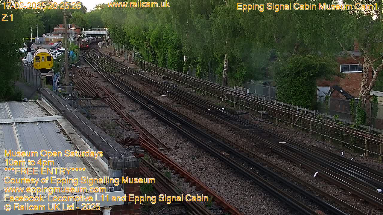 A high-angle, long shot of a railway scene at dusk, showing multiple train tracks, a train in the distance, and a yellow train car closer to the viewer.