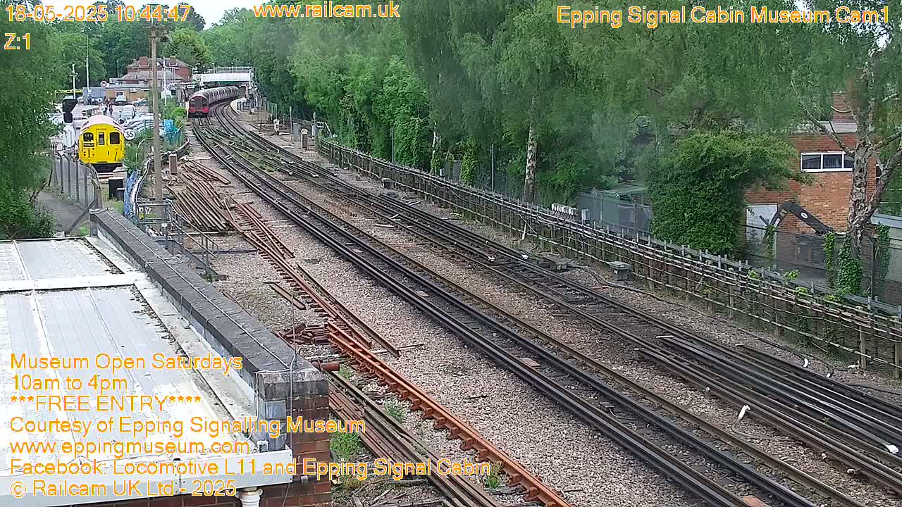 A high-angle view of a railway yard with multiple train tracks on a sunny day, featuring two trains, one yellow and one dark-colored, and lush green vegetation alongside the tracks.