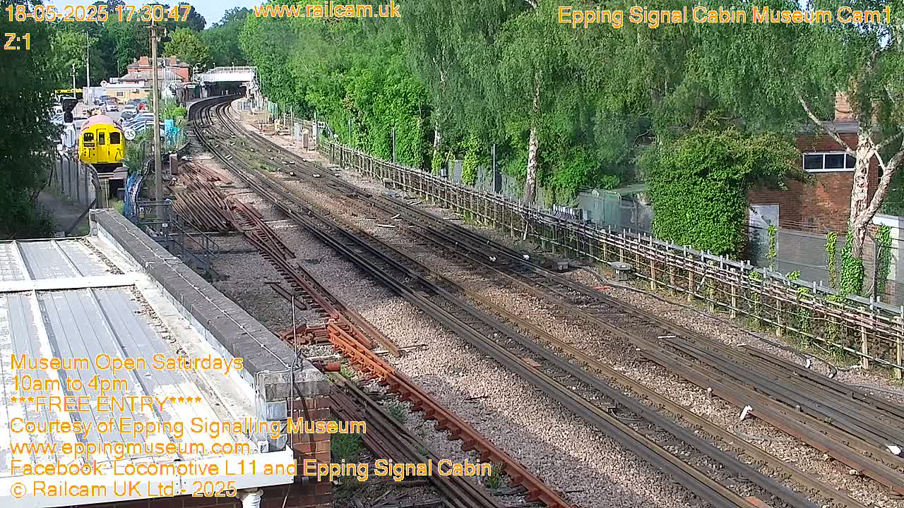 A yellow train is visible on a railway track, surrounded by lush green vegetation under a sunny sky.