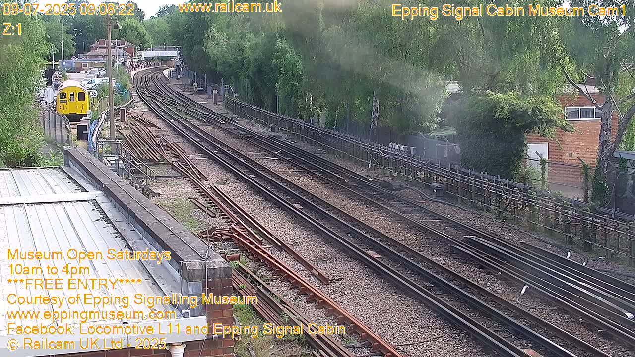 A high-angle, long shot shows multiple train tracks on a sunny day, with a yellow train car and a dark train engine visible.