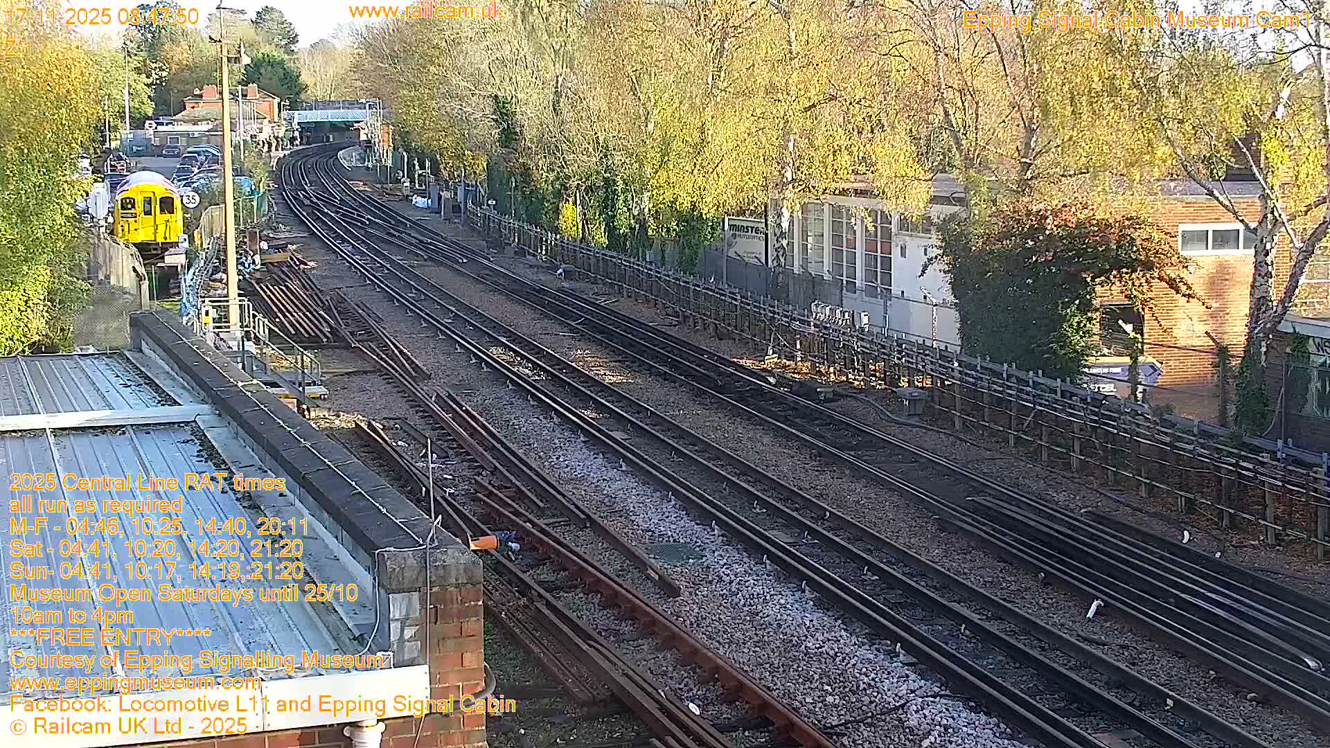 A high-angle view captures multiple railway tracks curving into the distance, bordered by buildings and trees with autumn foliage, under bright, sunny conditions, with a yellow train carriage visible on the far left.