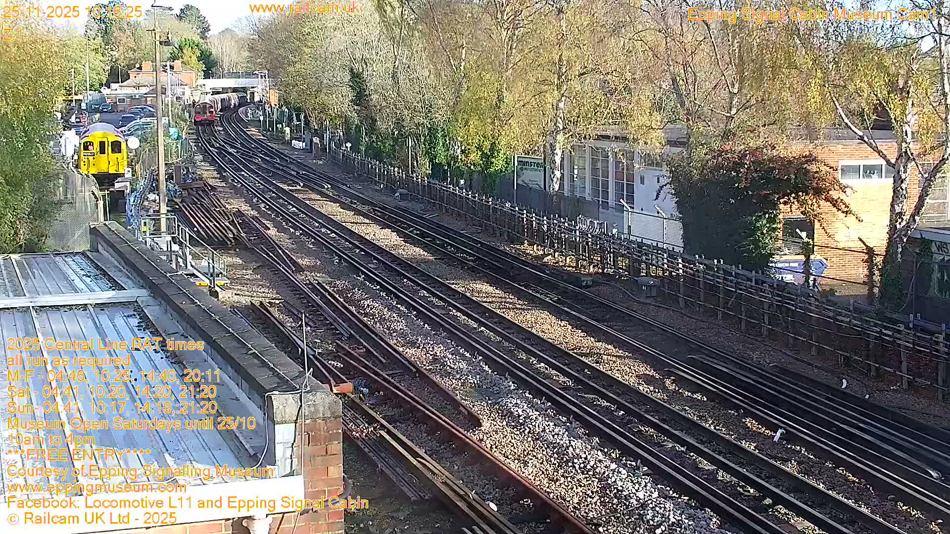 On a clear and sunny day, a network of railway tracks curves into the distance, featuring a yellow train on the left and a red train further along the line, bordered by buildings and deciduous trees with autumn leaves.