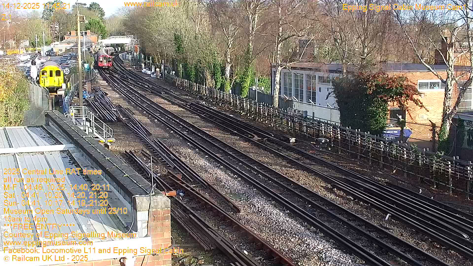On a clear and sunny day, a network of railway tracks curves into the distance, featuring a yellow train on the left and a red train further along the line, bordered by buildings and deciduous trees with autumn leaves.