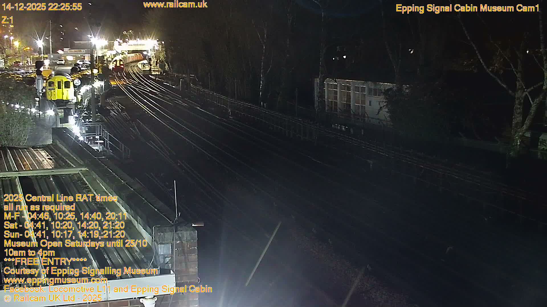 On a clear and sunny day, a network of railway tracks curves into the distance, featuring a yellow train on the left and a red train further along the line, bordered by buildings and deciduous trees with autumn leaves.