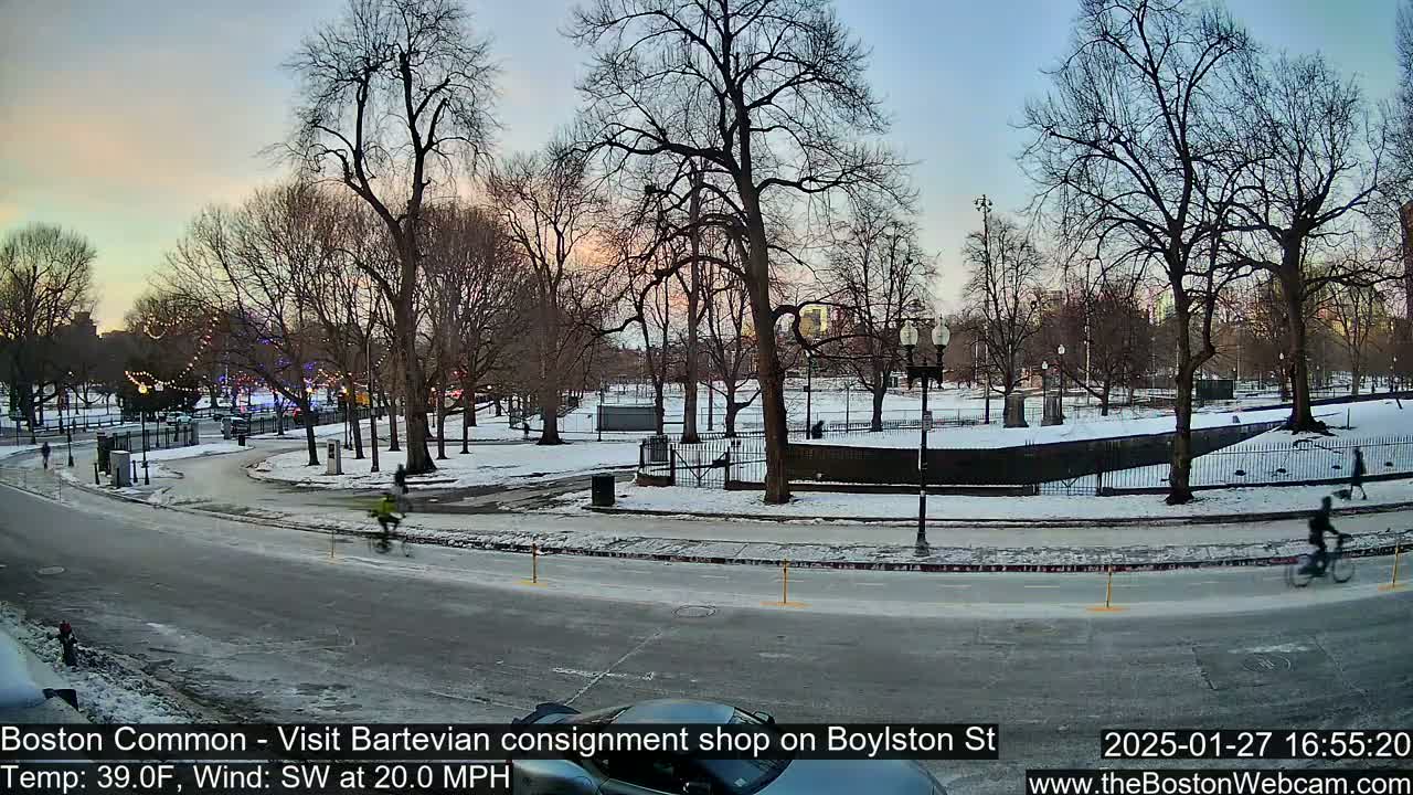 A snow-covered park with bare trees is visible from a street, where a few cyclists are riding past, under a twilight sky with a  southwesterly wind blowing at 20 mph.