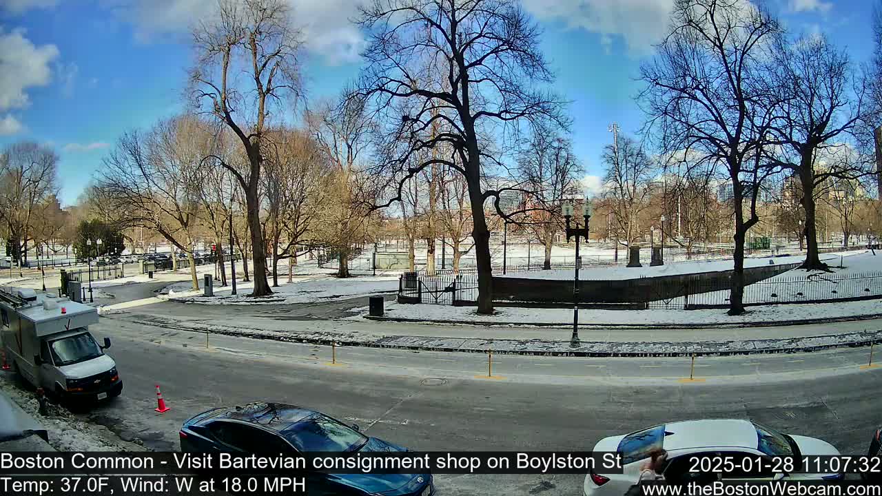A partly sunny winter day shows a snow-covered park and street with bare trees, a few cars, and a van.