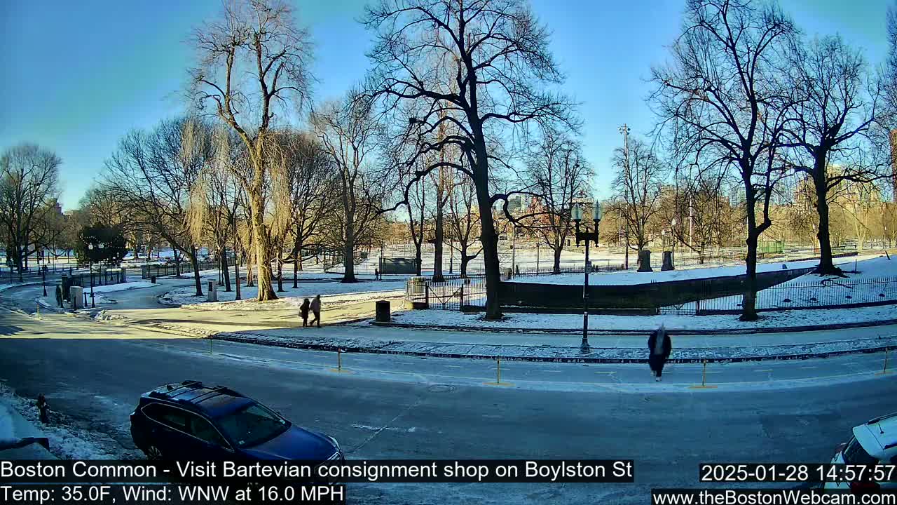 A partly sunny, cold day in a park with bare trees, some snow on the ground, and a few pedestrians walking near a road with cars parked.