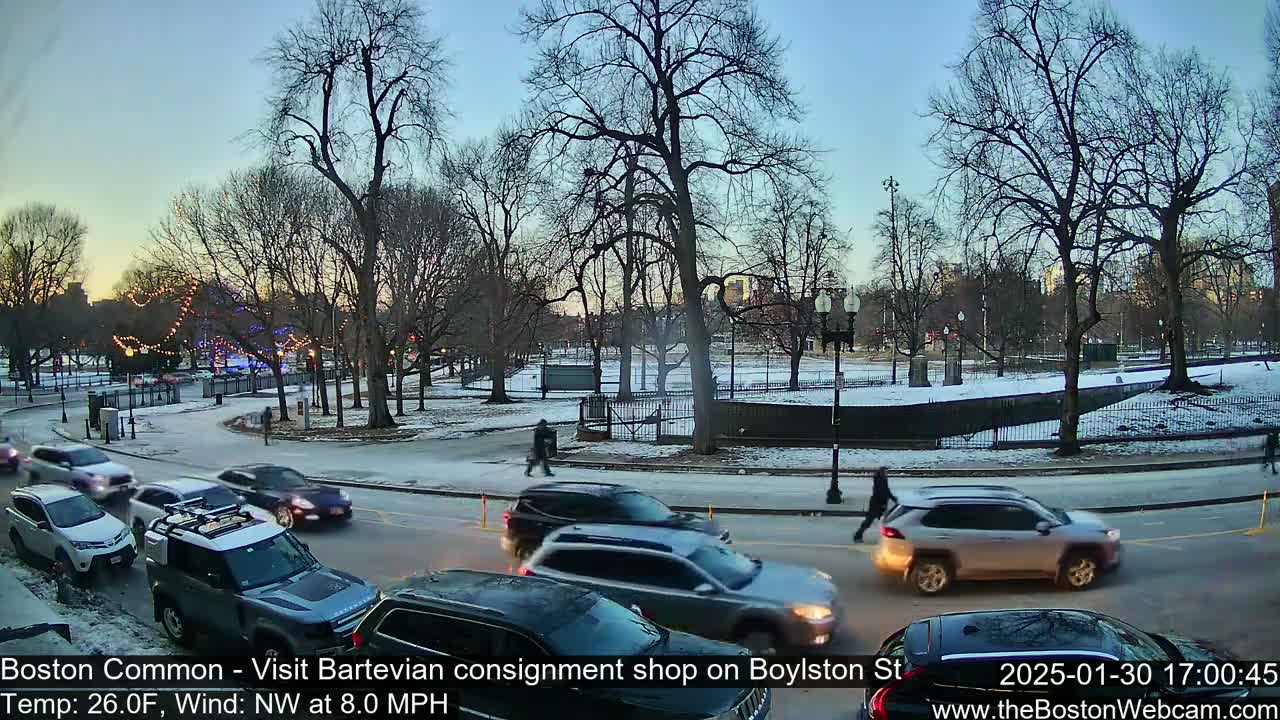 Cars drive along a snow-covered road near a park with bare trees under a twilight sky.