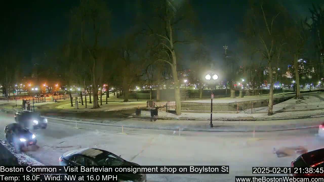 A nighttime scene of a snow-covered park with cars driving on a road, under a northwest wind blowing at 16 mph and a temperature of 18 degrees Fahrenheit.