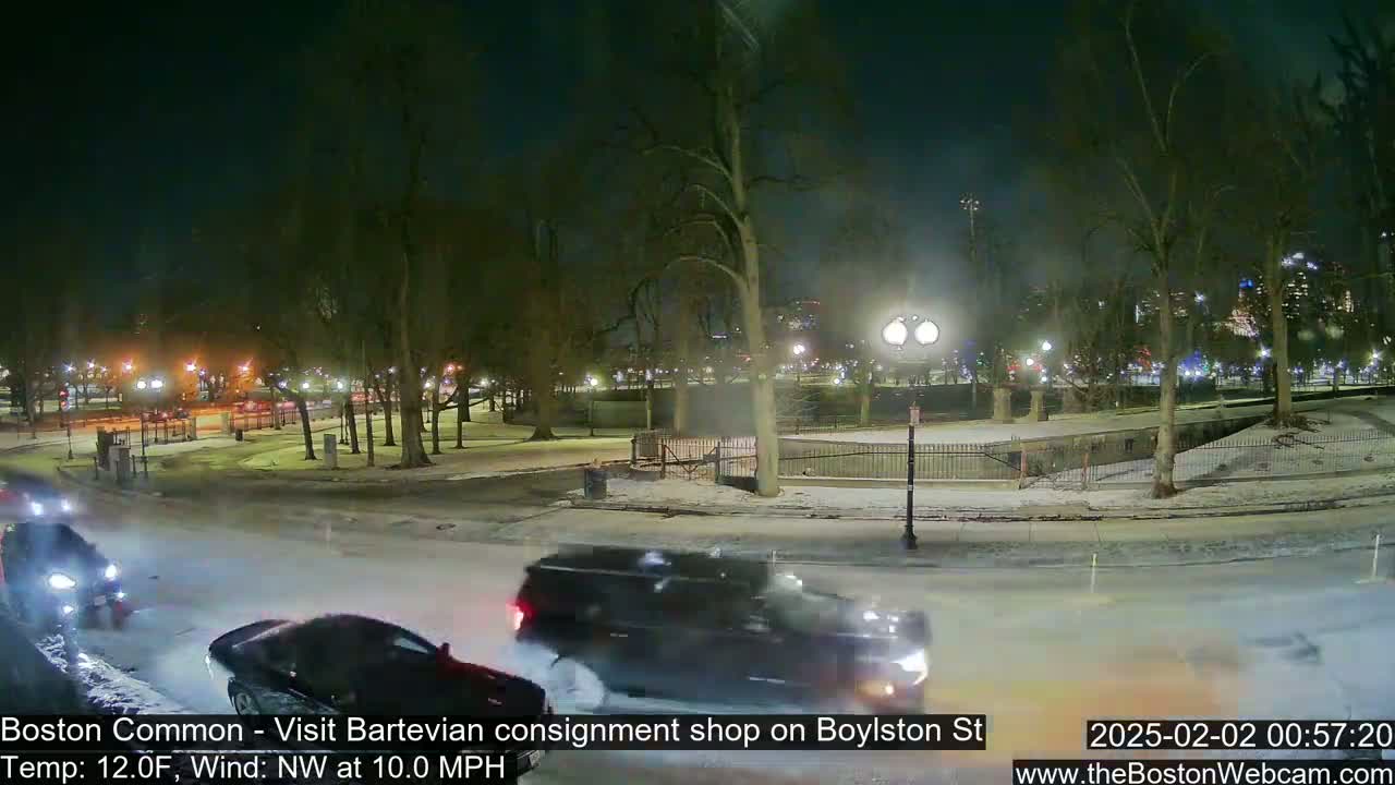 A snow-covered park at night with several cars driving on a nearby street under a dark sky, with a northwest wind blowing at 10 mph and a temperature of 12 degrees Fahrenheit.