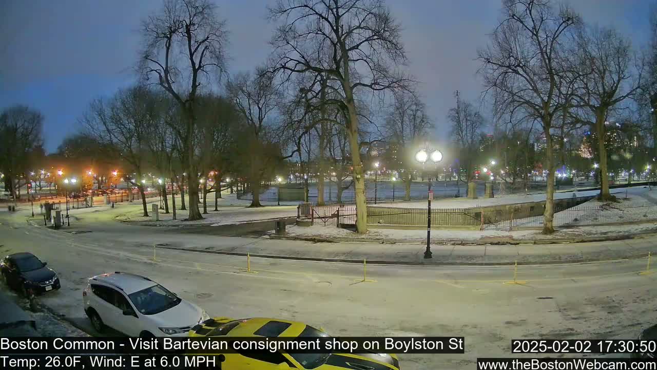 A snow-covered park at night, with cars parked along a street, is lit by streetlights under a twilight sky.