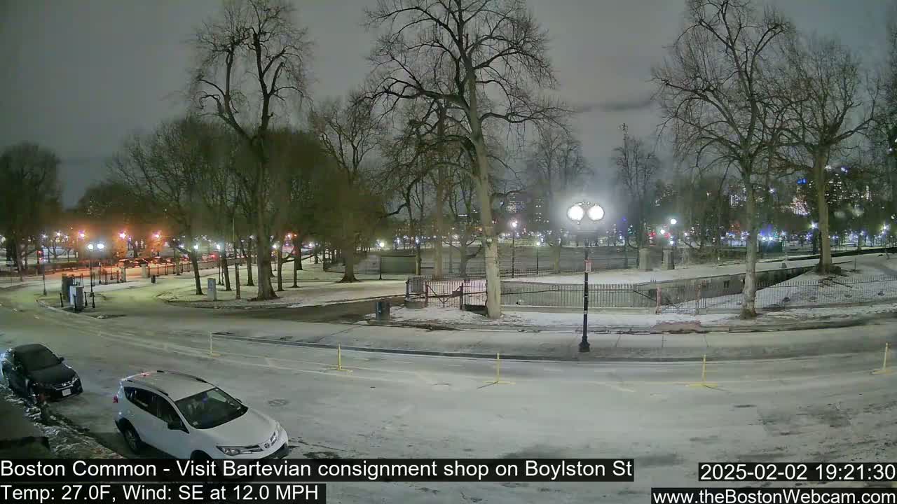 A snow-covered park at night, with a few cars parked along a road, under a cloudy sky, and with a wind blowing from the southeast at 12 mph.