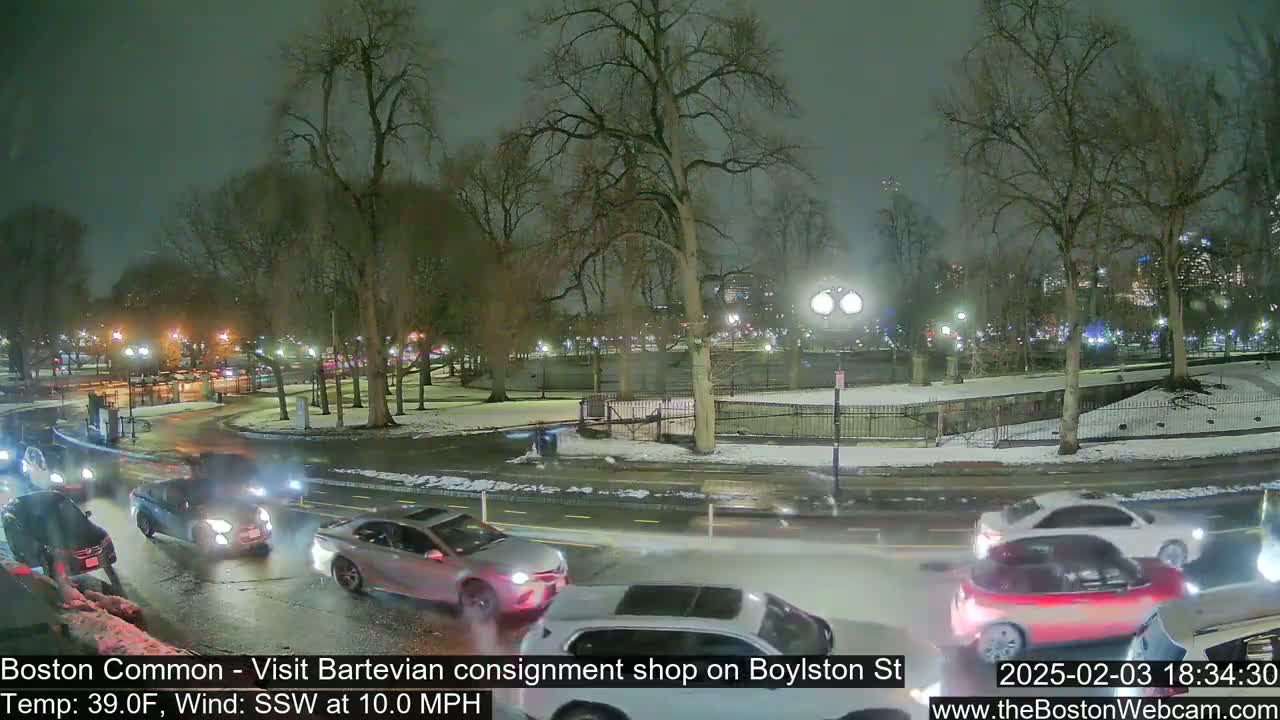 A nighttime view of a snow-dusted park with several cars driving along a road bordering it, under a cloudy sky.