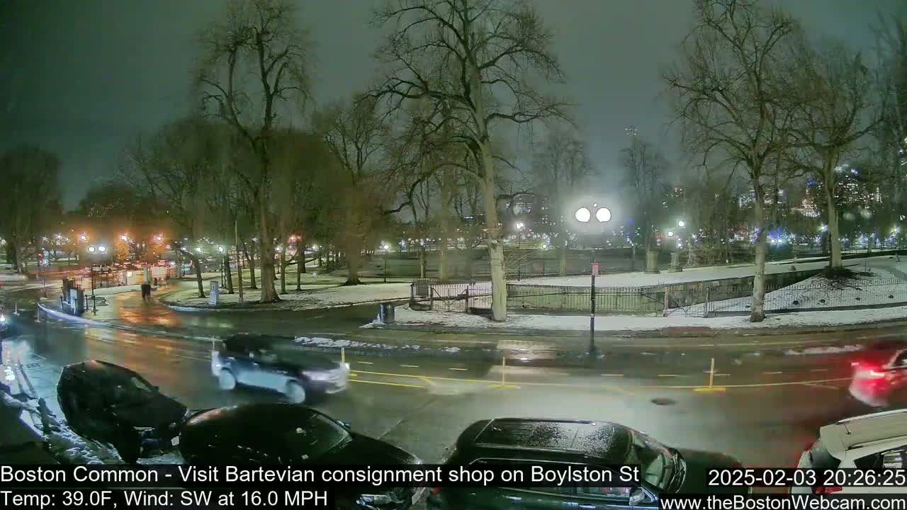 A nighttime view of a snow-dusted park with cars driving on a wet road, under a dark, overcast sky.