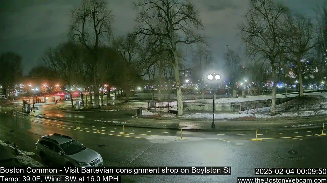 A nighttime view of a park with wet roads and some snow on the ground, under a dark sky, with a vehicle parked near the edge of the road.