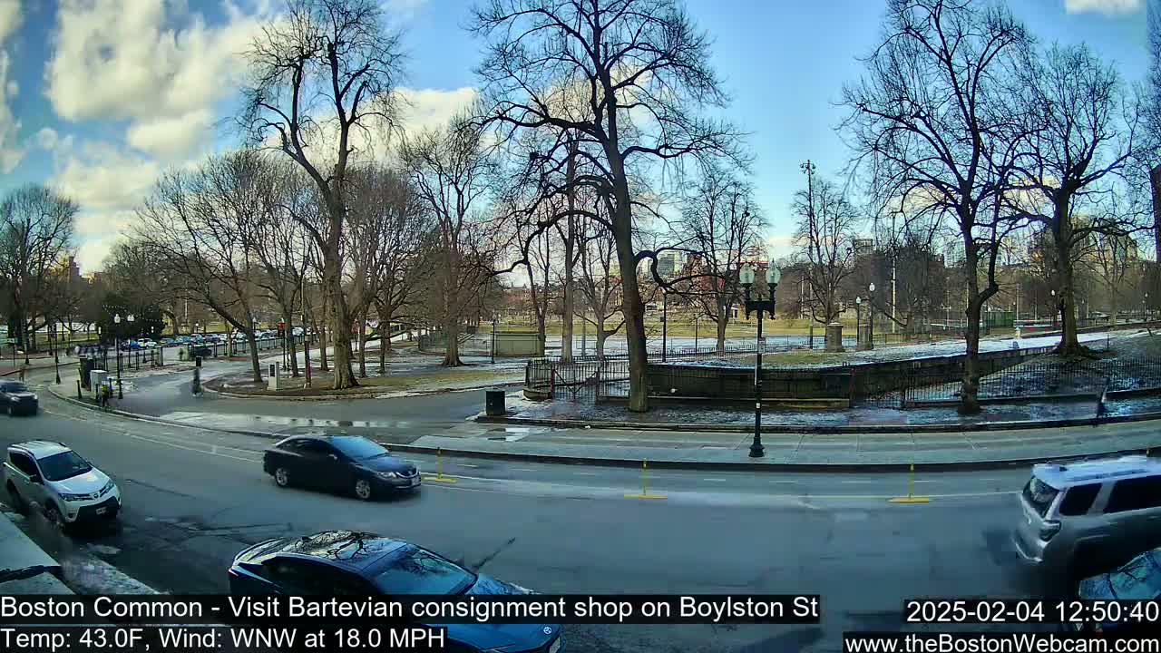 A partly cloudy day with a wind of 18 mph shows a street curving around a park with bare trees, several cars driving on the road, and some snow and ice on the ground.
