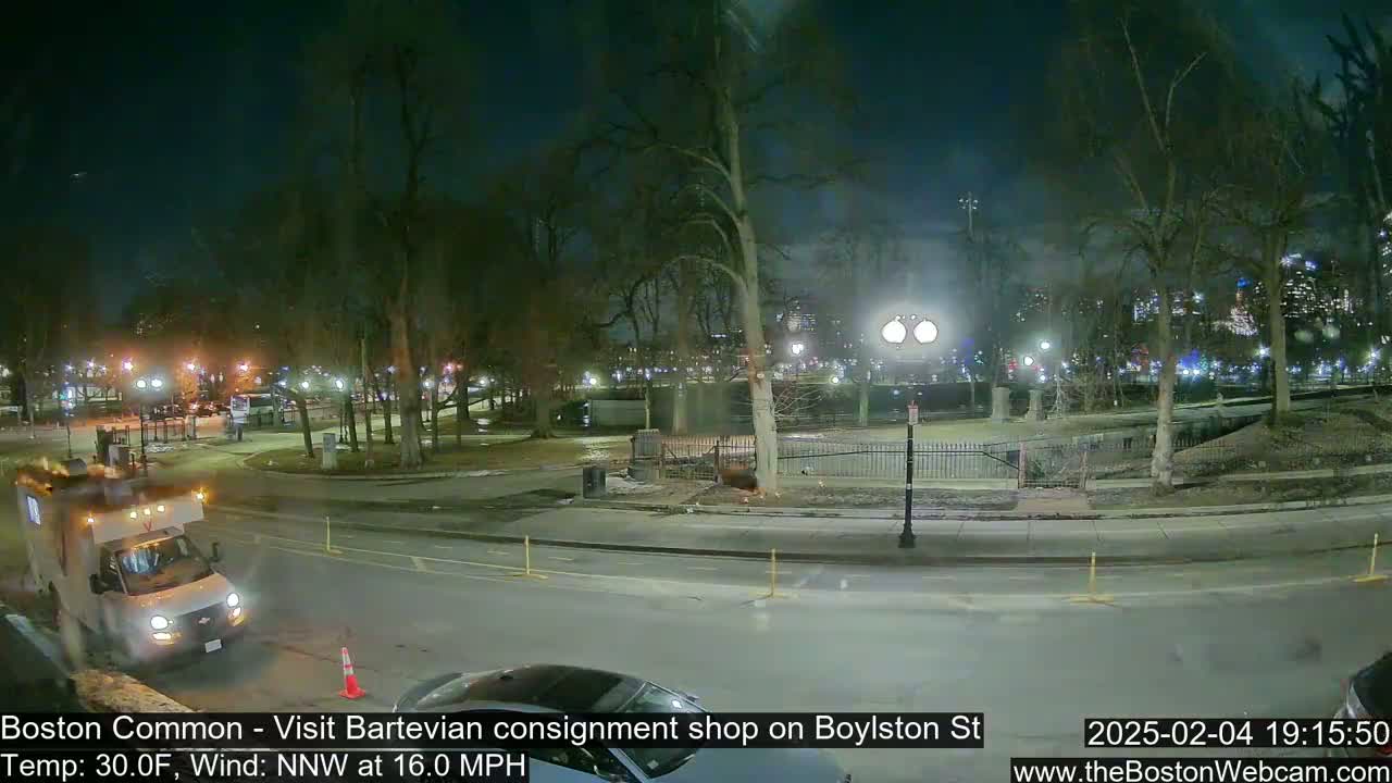 A nighttime street scene in a park, with a van parked on the road, bare trees, and city lights in the background, under a windy, 30°F temperature.