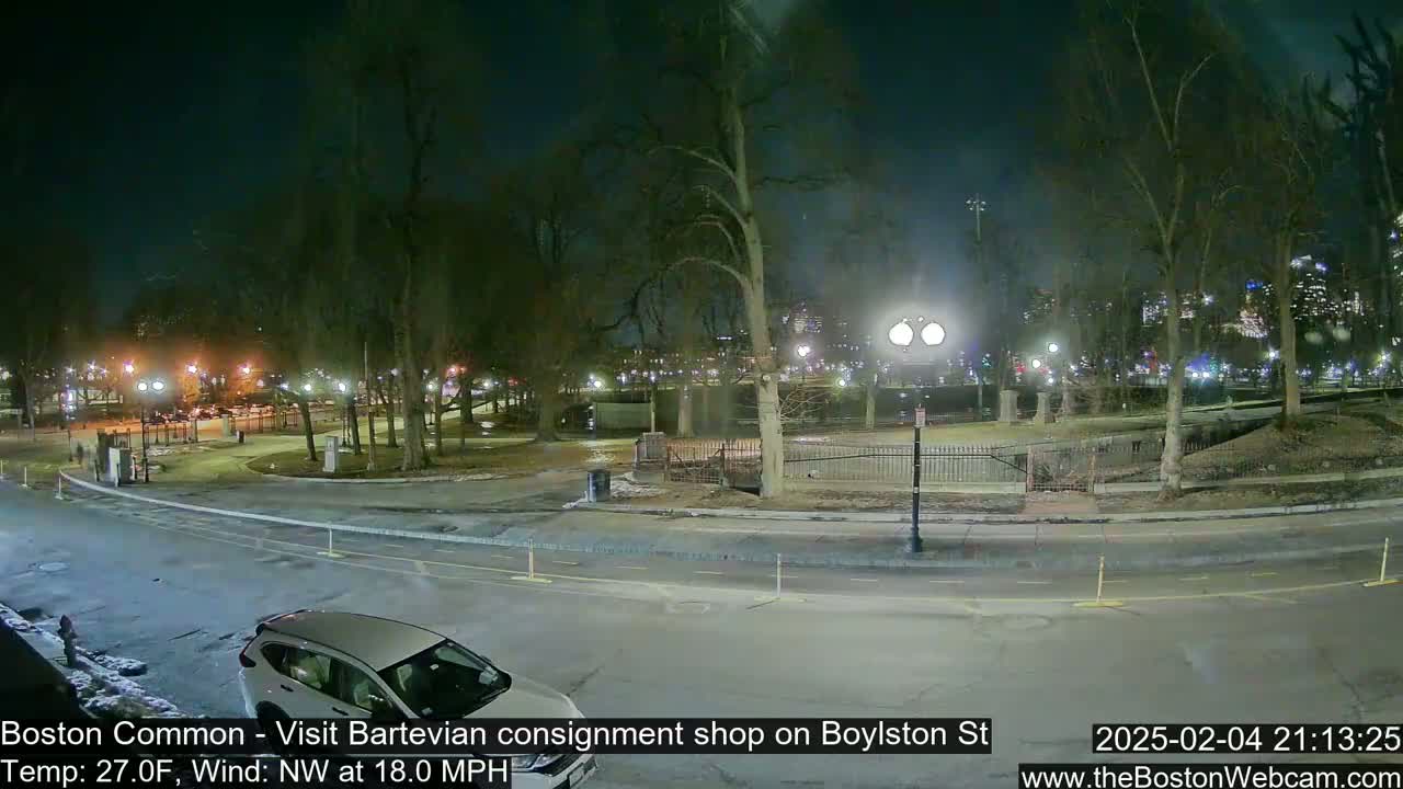 A nighttime view of a park with bare trees, streetlights, and a car parked near a roadway, under a windy, 27°F weather condition.