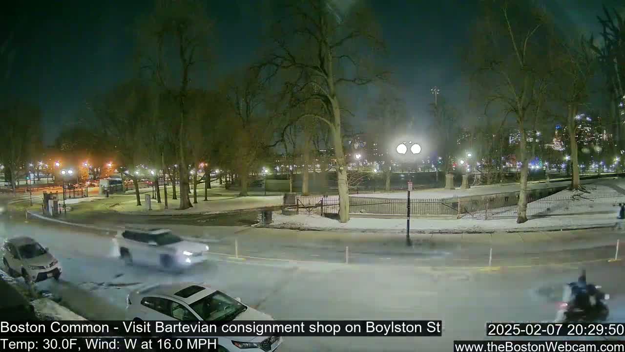 A nighttime view of a snow-covered park and street with several cars driving by under a dark sky with a westerly wind.