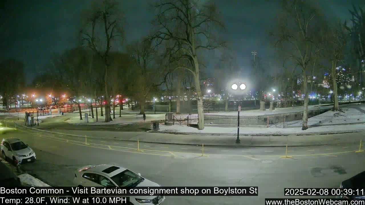 A nighttime view of a snow-dusted park with several cars parked along a street, under a dark sky.