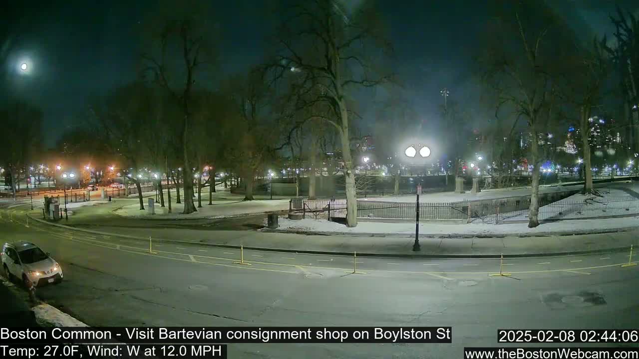 A nighttime view of a snow-covered park with a street and a car, under a partially cloudy sky with a visible moon.