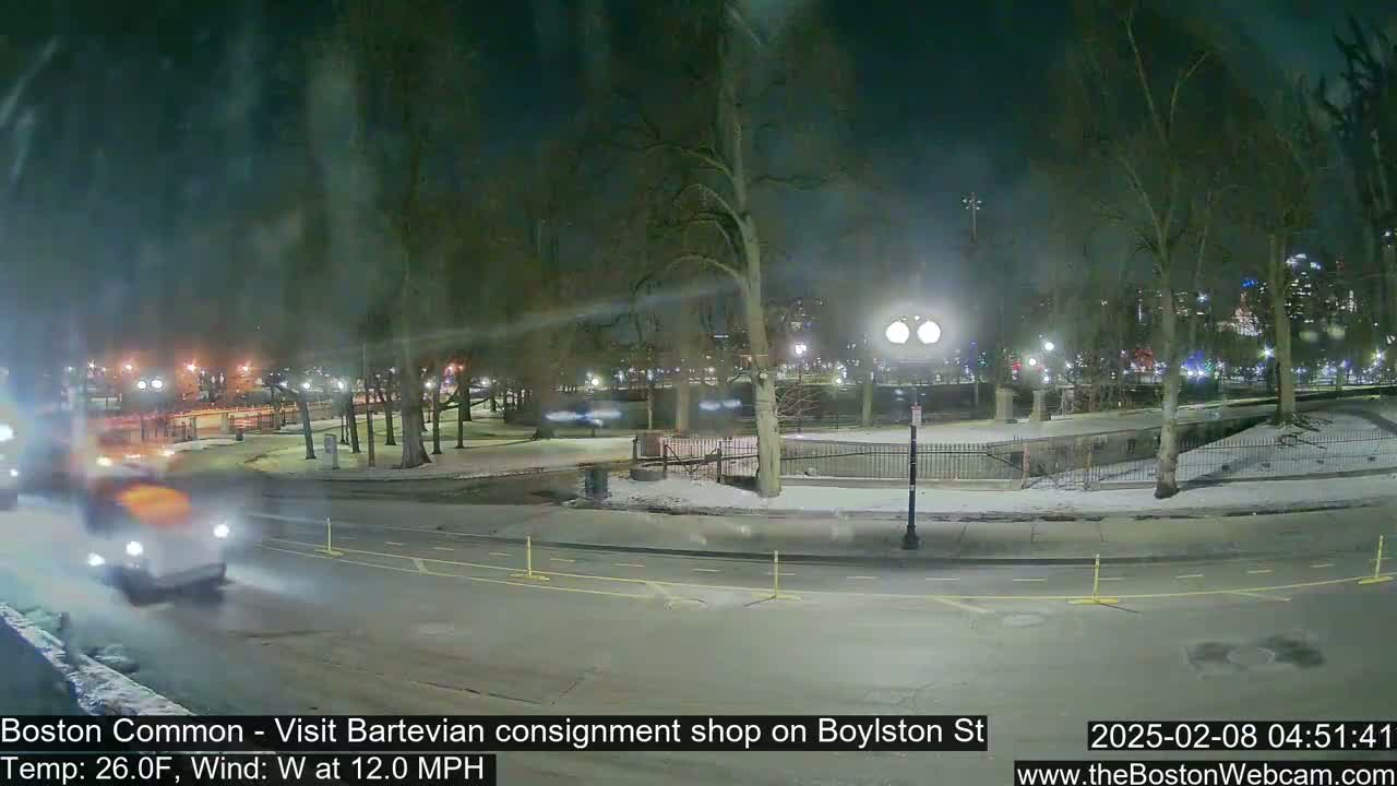 A nighttime view of a snow-covered park with a road and cars passing by under a windy, 26°F weather condition.