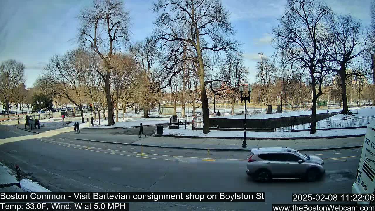 A city park with bare trees and patches of snow is visible from a street, with a few pedestrians and a silver SUV driving past.