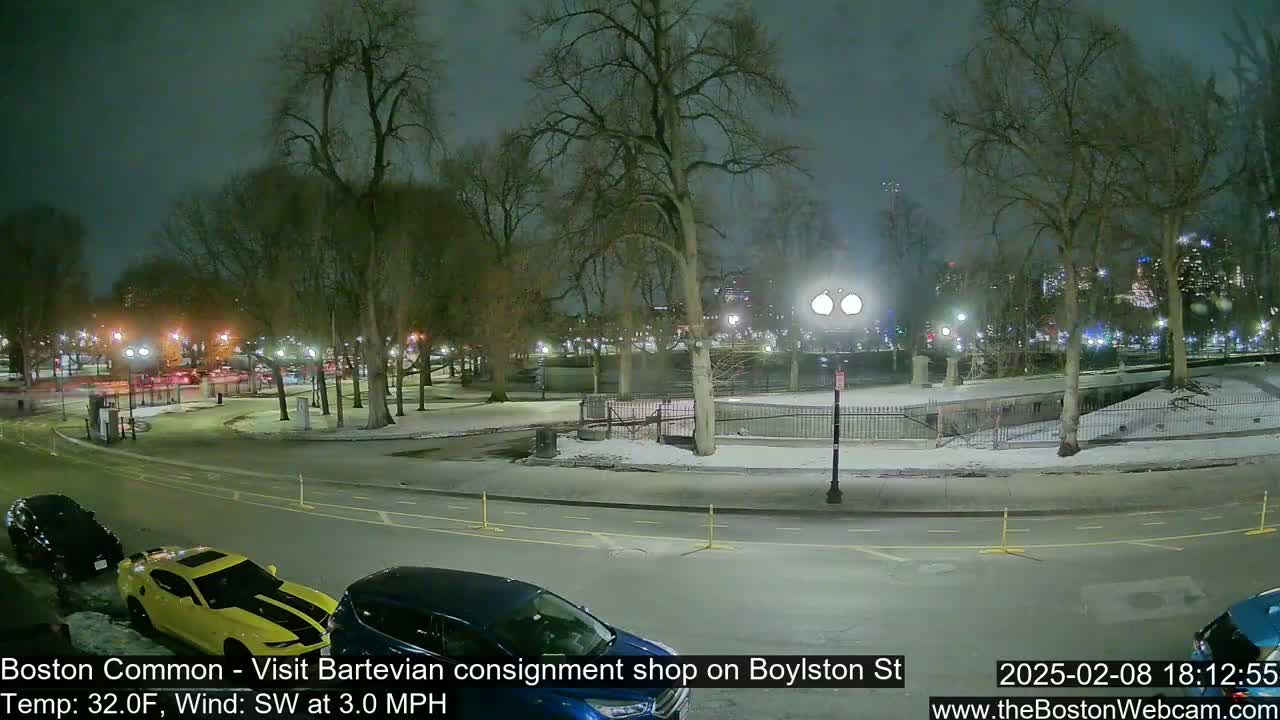 A nighttime view of a snow-covered park with cars parked along a street, under a cloudy sky.