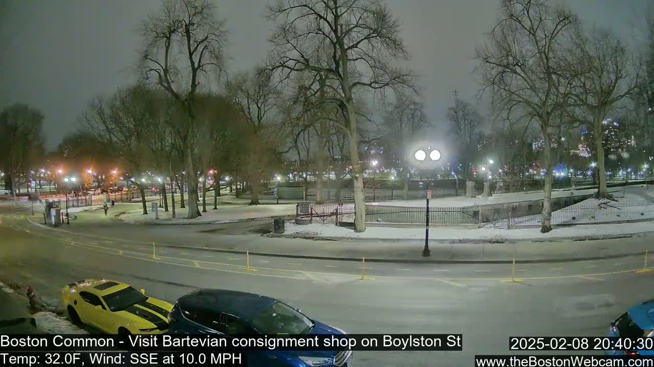 A nighttime view of a snow-dusted park with a few cars parked near a road, under a cloudy sky.