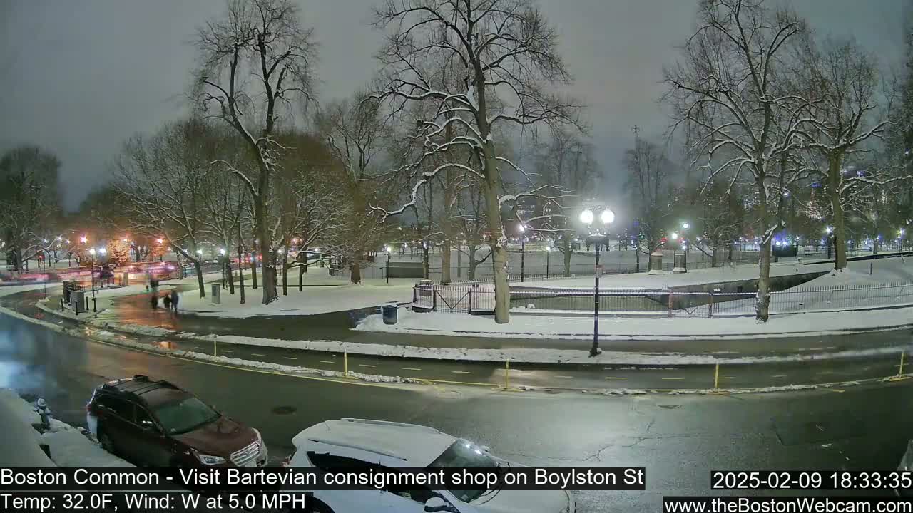 A snow-covered park at night, with a few people walking and cars parked along a nearby road.