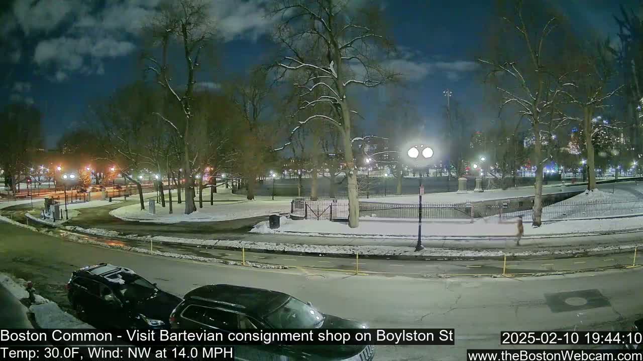 A snow-covered park at night with a few cars parked nearby under a partly cloudy sky.